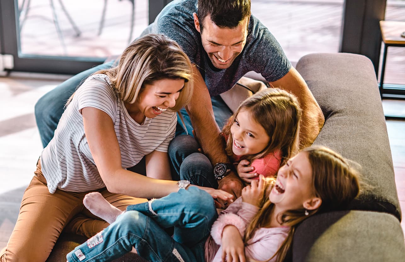 familia, papá y mamá haciendo cosquillas y dos hijas sobre el sofá del living